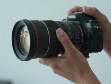 A close-up shot of a professional photographer's hand adjusting a high-end lens. The lighting is soft and cinematic, with a palette of light blue and dark gray. Modern studio environment in Latin America.