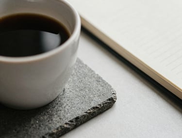 Close-up of a cup of coffee on a grey stone coaster next to a closed notebook, neutral colors, representing a quiet moment of introspection.