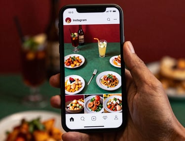 Hand holding a smartphone displaying an Instagram feed full of vibrant food photos in Deep Crimson Red and Matte Forest Green tones. European / Iberian setting.