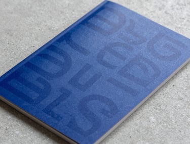 Close-up photography of a high-quality printed marketing brochure featuring bold typography and deep dark blue colors, lying on a light grey concrete surface.
