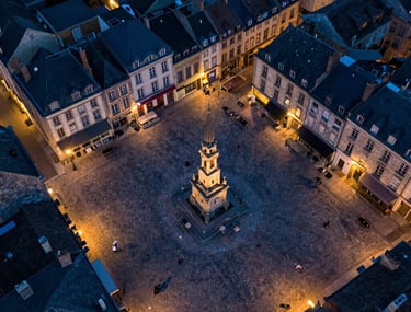 Drone view of a historic French town square at dusk. The warm street lights contrast with the deep dark blue of the cobblestones.