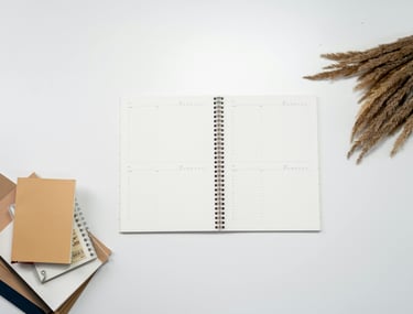 Flat lay of an open spiral notebook planner on a white desk with stacked journals and dried pampas grass.