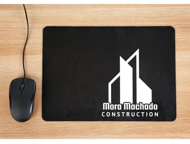 Black mouse pad with white Mora Machado Construction logo next to a computer mouse on a wood desk.
