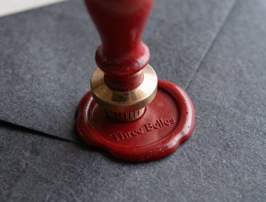 Macro shot of an antique wax seal in blood red being pressed onto a charcoal gray envelope, symbolizing the premium quality of Three Belles' work.