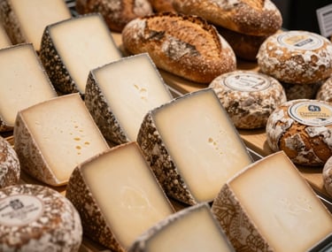 Close-up overhead shot of a modern food market display, showcasing artisanal cheeses and rustic breads under warm, focused lighting in a Northern European / North American setting.