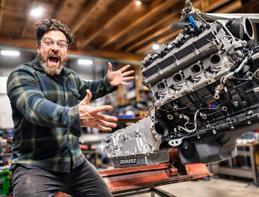 Excited mechanic in a garage posing with a powerful car engine suspended on a hoist.
