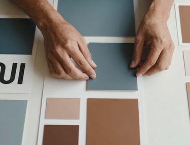 Top-down view of a designer's hands arranging minimalist color palettes and typography samples on a clean desk. Latin American / Hispanic context, professional lighting, muted steel blue and warm bronze colors visible.