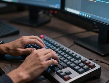 A close-up of a developer's hands typing on a mechanical keyboard. The scene is lit by the glow of monitors in a dark room with slate gray blue accents. Professional Latin American setting.