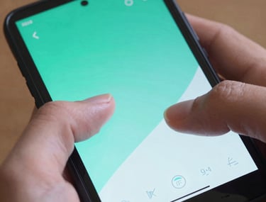 Detailed photograph of a user's thumb interacting with a beautifully designed Android interface on a handheld device. The screen displays vibrant teal and light blue gradients. North American / US setting, professional lighting.