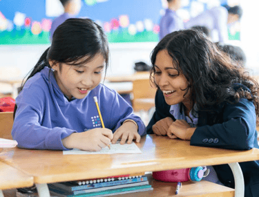 A female teacher  to the right of a student, teacher her with a smile on her face. The student is working happily, writing.