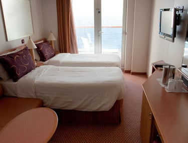 Cruise-ship cabin with two twin beds, a balcony door overlooking the ocean, wall-mounted TV, small sofa, and a desk