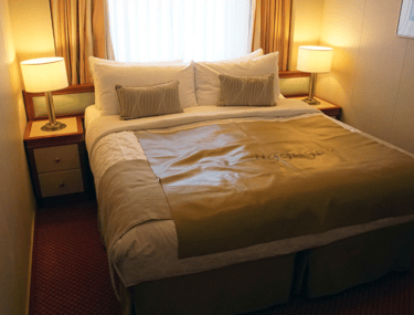 Cozy cruise-ship stateroom with a king bed, two bedside tables and lamps, and a curtained window above the headboard