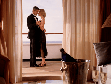 Formal couple dancing on a cruise-suite balcony with the ocean beyond, seen through curtains, with champagne and flutes