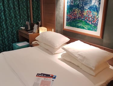 Cozy cruise-ship stateroom with a made king bed and stacked pillows, bedside lamps, green patterned curtains, a framed painti