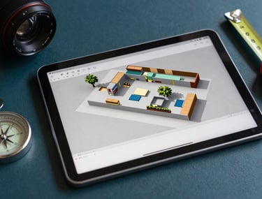 A professional photograph of an art director's kit, featuring a digital tablet showing a 3D set model, a compass, and metal measuring tapes on a dark teal surface. The lighting is crisp and professional.