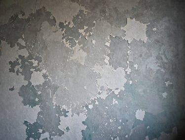A detailed close-up of a weathered North American interior wall on a film set. The surface shows layers of peeling silver-grey paint and dark teal undertones, masterfully aged by a scenic artist. Soft, moody side-lighting highlights the texture and depth of the plaster work.