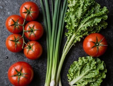Top-down photography of a chef's workstation with various fresh, colorful ingredients like ripe tomatoes and vibrant greens on a dark matte surface.