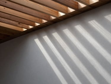 Sunlight streaming through slats in a wooden ceiling, creating a pattern on a light gray wall. High-contrast architectural detail.