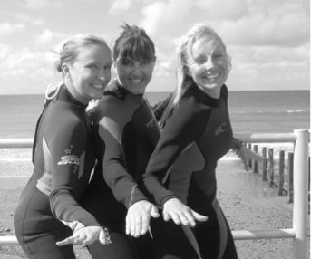 3 women in wetsuits posing in surfer stances next to the beach.