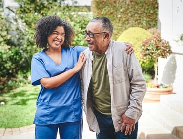 A nurse stands beside an elderly man outside, both smiling and engaged in conversation in a sunny ou