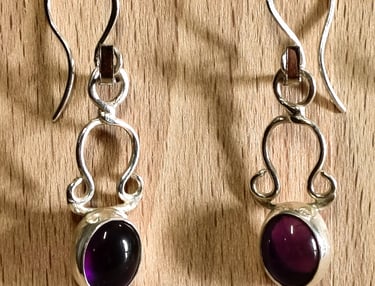 Handmade sterling silver drop earrings with oval purple amethyst gemstones on a wooden background.