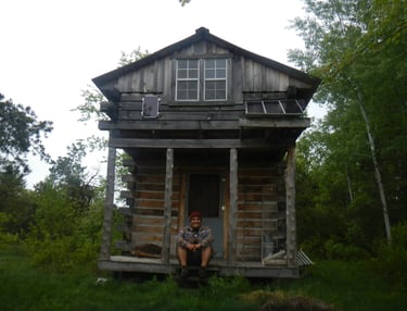 a smiling person sits on the front porch of a rustic wood cabin