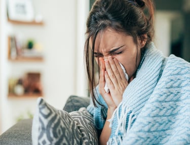 A woman sneezing into a tissue while wrapped in a blue blanket, suffering from cold or allergy symptoms.