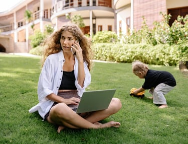 Woman working remotely on laptop and phone in a park while a child plays, symbolizing flexib