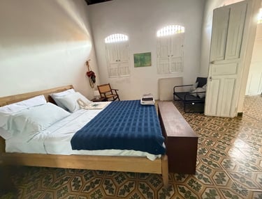 Rustic bedroom with blue bedspread, wooden bed frame, patterned tile floor, and white colonial shutters.