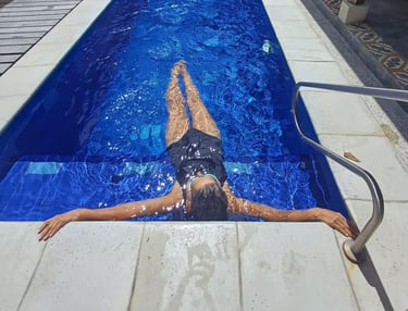A woman in a black swimsuit relaxes in a blue backyard swimming pool with arms outstretched.