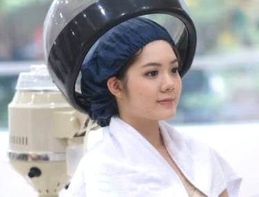 A young woman with a hair bonnet sitting under a professional salon hooded hair dryer for a treatment.