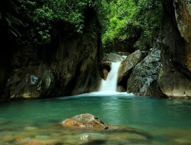 Kolam air terjun alami dengan air berwarna hijau toska yang jernih, dikelilingi tebing batu gelap dan vegetasi hutan hijau le