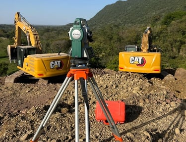 Sokkia total station on a tripod at a construction site with two CAT excavators and hills.