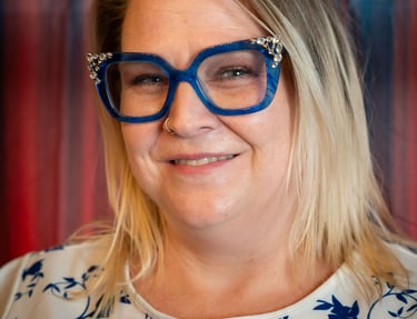 Woman with blonde hair wearing stylish blue floral cat-eye glasses and a white floral blouse.