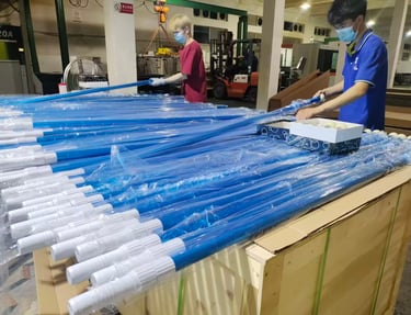 Factory workers packing blue telescopic pool cleaning poles into protective plastic sleeves for shipping.