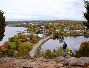 A man looks out from a rocky overlook at a scenic waterfront town with autumn foliage.