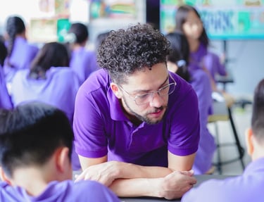 Male teacher leaned overlooking at a students work. In front of the teacher are two students leaned over doing their work.