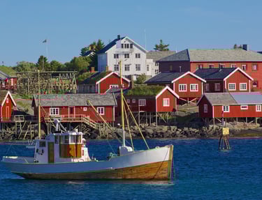 Fiskebåt foran røde rorbuer i Lofoten, sjø og bryggeanlegg i sollys.