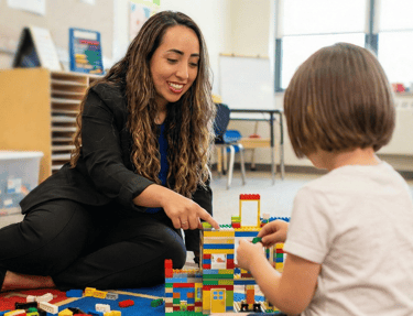 Julie Granata sorri enquanto realiza atividade lúdica com blocos de montar com uma criança em ambiente acolhedor