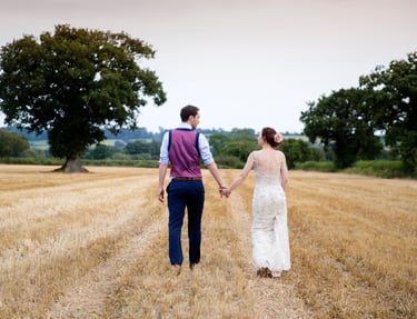 A bride and groom holding hands while walking through a golden rural field after their wedding ceremony.