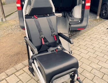 Mobilitätsstuhl mit roten Gurten auf Rampe hinter grauem Fahrzeug, draußen.