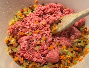 Browning raw ground beef with sautéed carrots, onions, and celery in a white pot for bolognese sauce.