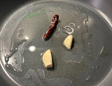 Fresh garlic cloves and a dried red chili pepper sizzling in olive oil inside a non-stick frying pan.