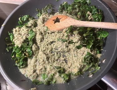 Sautéed green leafy vegetables and creamy pesto sauce cooking in a black frying pan with a wooden spoon.