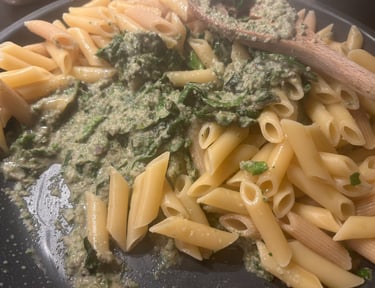 Cooked penne pasta being tossed with creamy green spinach pesto sauce in a large skillet.