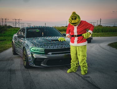 The Grinch in a Santa suit stands next to a gray Dodge Charger wrapped in festive Christmas lights at sunset.