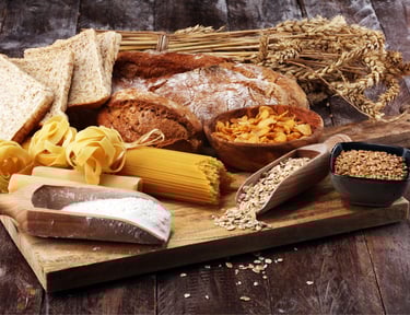 An assortment of whole grain foods including rustic bread, pasta, oats, and wheat stalks on a wooden board.