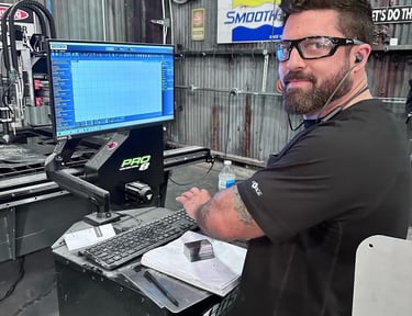 A bearded technician wearing safety glasses operates a CNC machine control console in a metal workshop.