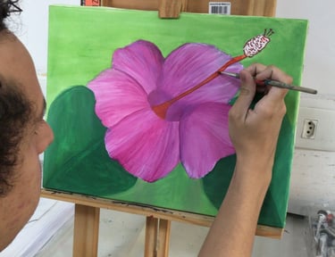 Artist painting a pink hibiscus flower on a green canvas using a fine brush and wooden easel.