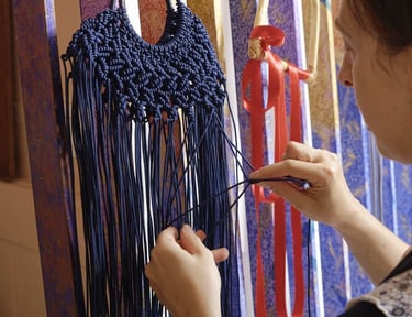 Manos tejiendo un bolso de macramé azul con flecos.”